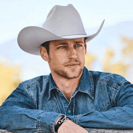 Hombre con sombrero vaquero blanco, camisa de mezclilla, fondo campestre.
